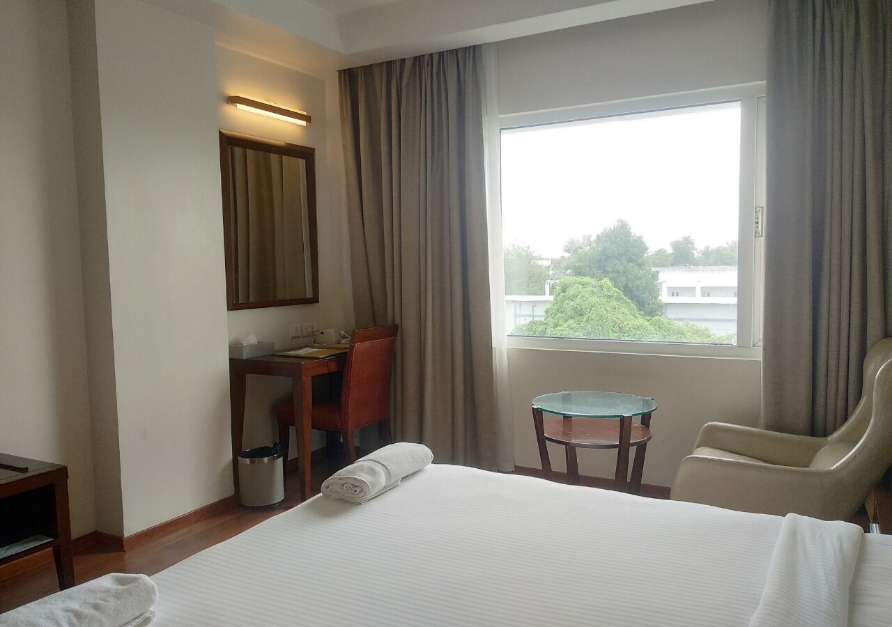 A hotel room with a large bed, a desk with a chair, a small table, and a window with curtains.