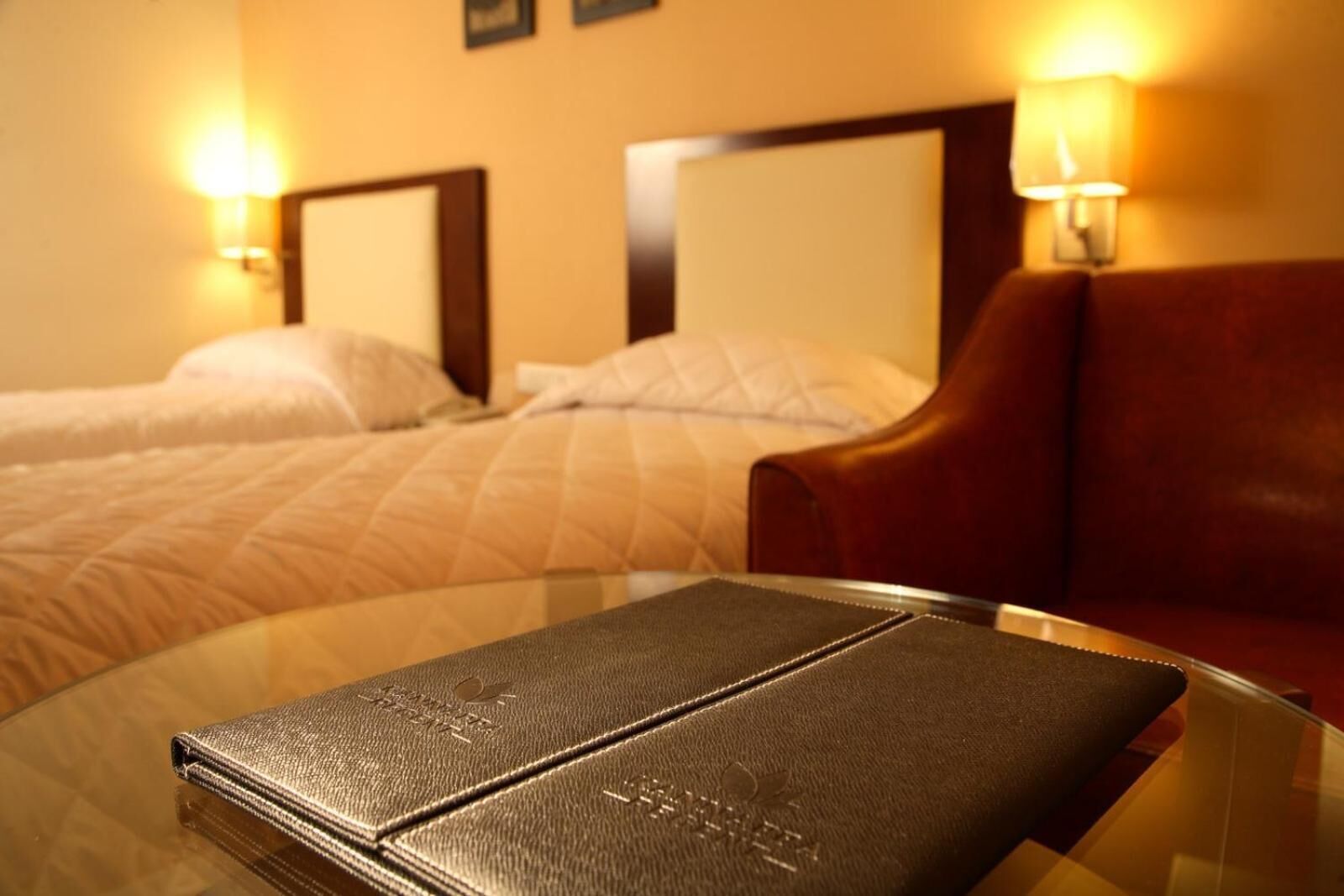 A hotel room with a bed, two bedside lamps, a leather armchair, and a glass table with a book.