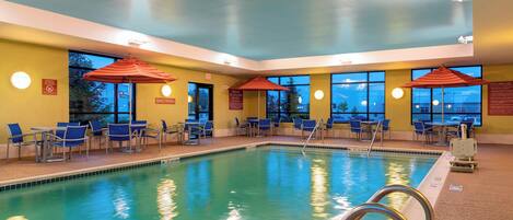An indoor swimming pool with blue chairs and red umbrellas, surrounded by yellow walls and large windows.