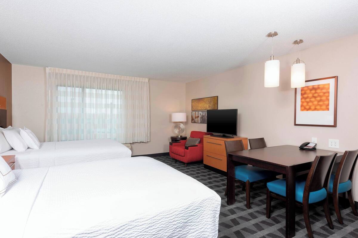 A hotel room with two beds, a desk with a chair, a television, and a dining table with chairs.