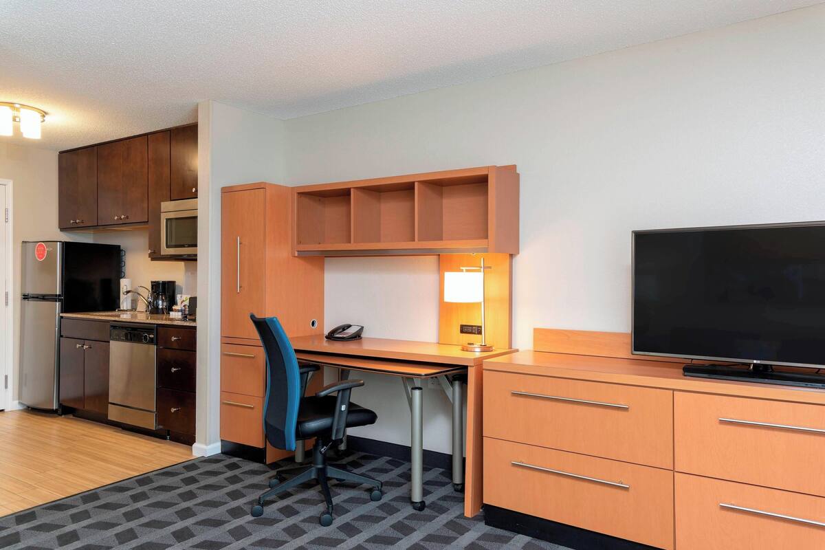 A compact hotel room with a kitchenette, a desk with a chair, a television, and a small cabinet.