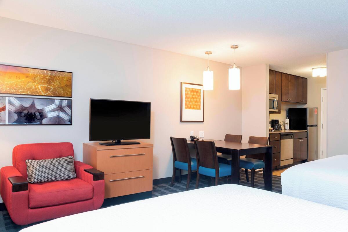 A hotel room with a bed, a TV, a desk with chairs, a kitchen area, and artwork on the walls.