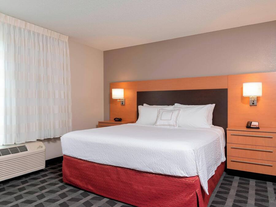 A hotel room with a large bed, two bedside tables with lamps, a window with curtains, and a wall-mounted air conditioner.