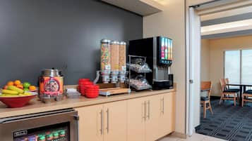 Eine Küchenzeile mit einem Kühlschrank, einer Kaffeemaschine und einer Mikrowelle. Außerdem befinden sich dort Snacks in Behältern und eine Schüssel Obst.