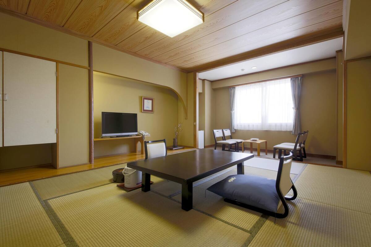 A traditional Japanese-style room with tatami flooring, a low table, chairs, a television, and a window with curtains.