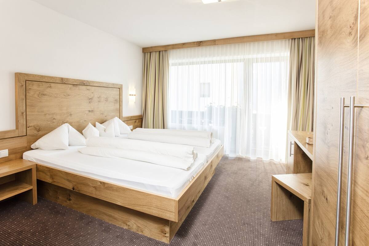 A hotel room with a large wooden bed, a wooden nightstand, a small wooden desk, and a large window with sheer curtains.