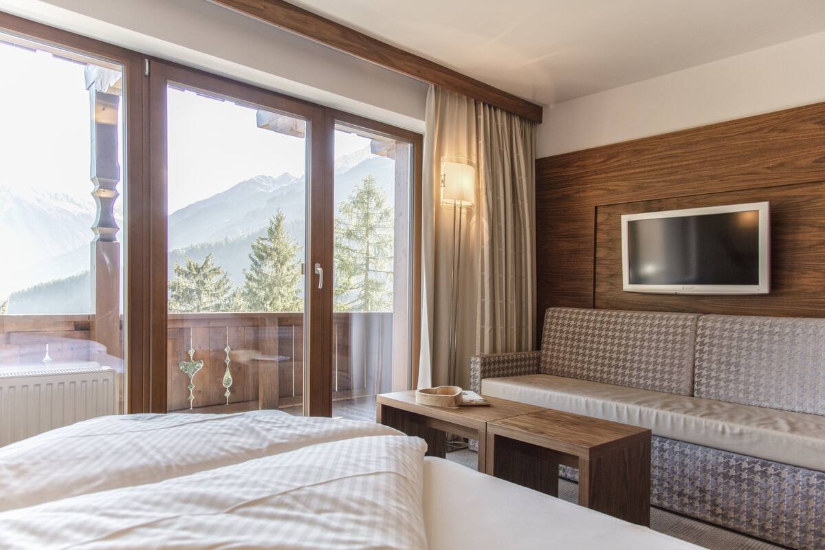 A hotel room with a bed, a sofa, a TV mounted on the wall, and a view of mountains through the window.