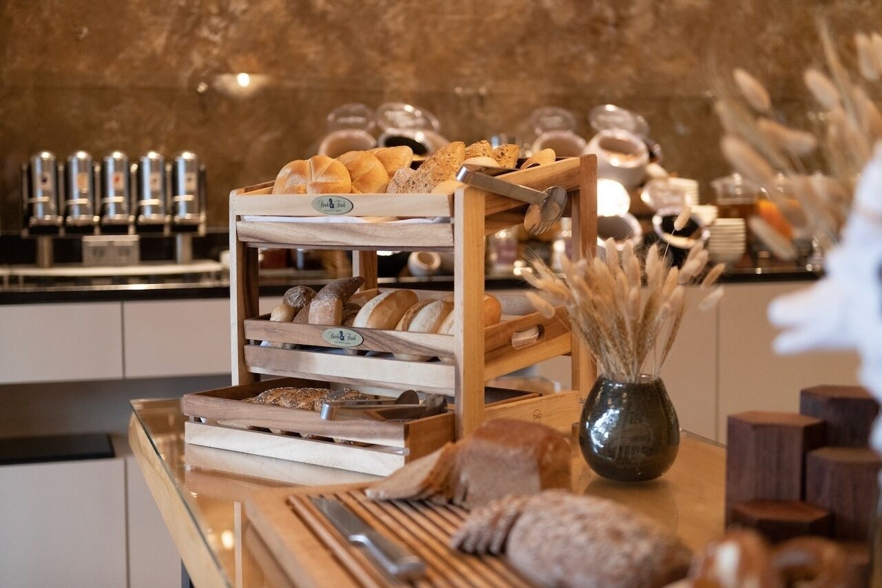 Una vitrina de panadería con diversos panes y pasteles, una máquina de café y un jarrón con plantas secas.