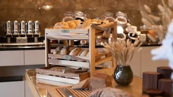Una vitrina de panadería con diversos panes y pasteles, una máquina de café y un jarrón con plantas secas.