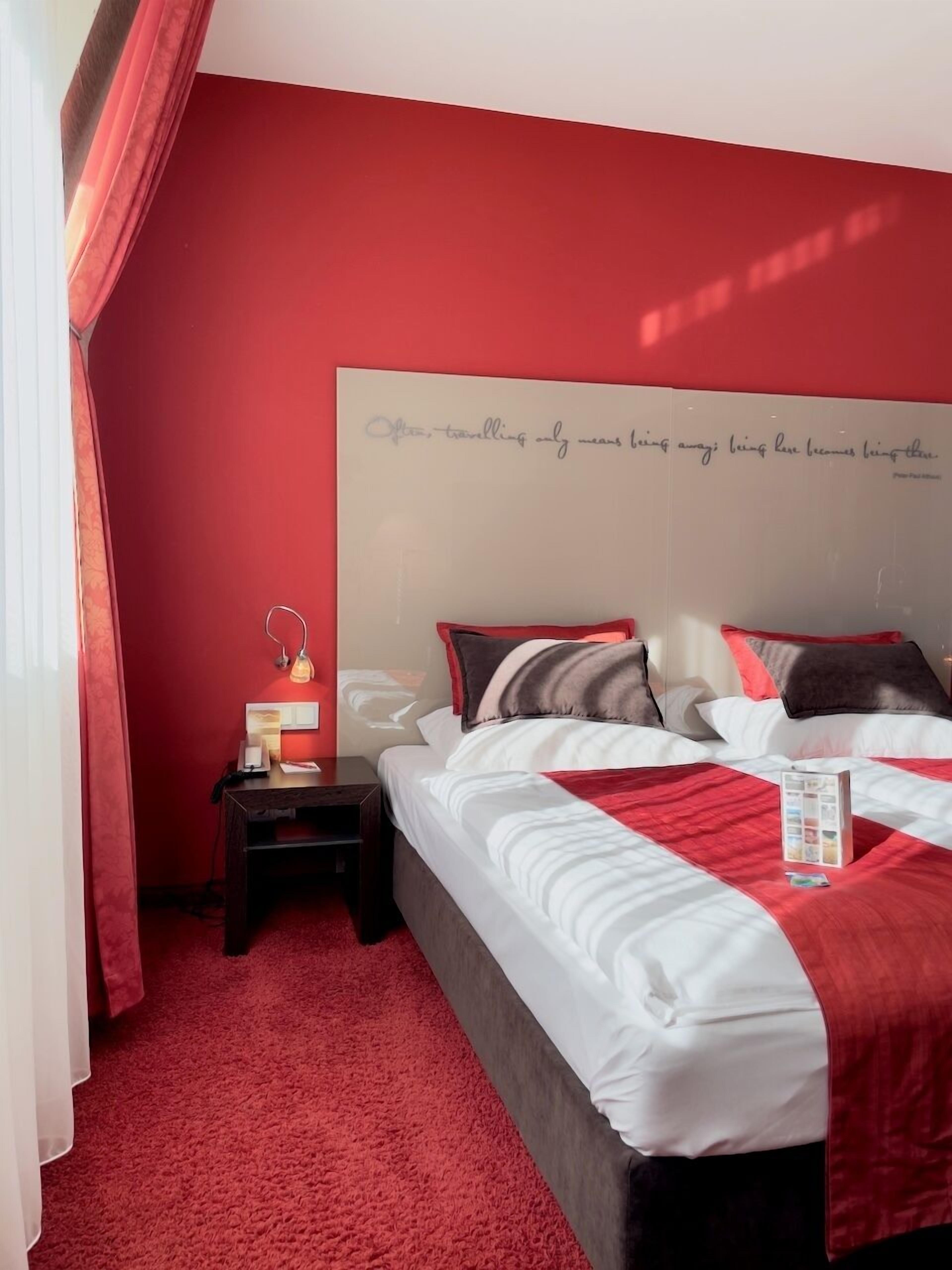 A neatly arranged bedroom with a red carpet, a bedside table with a lamp, and a bed with a red and white bedspread.