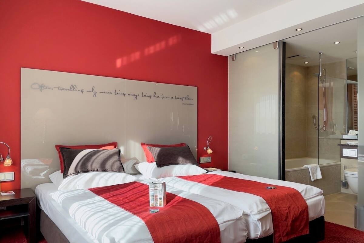 A modern hotel room with a large bed, a glass shower enclosure, and a red accent wall.