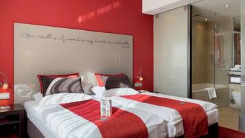 A modern hotel room with a large bed, a glass shower enclosure, and a red accent wall.