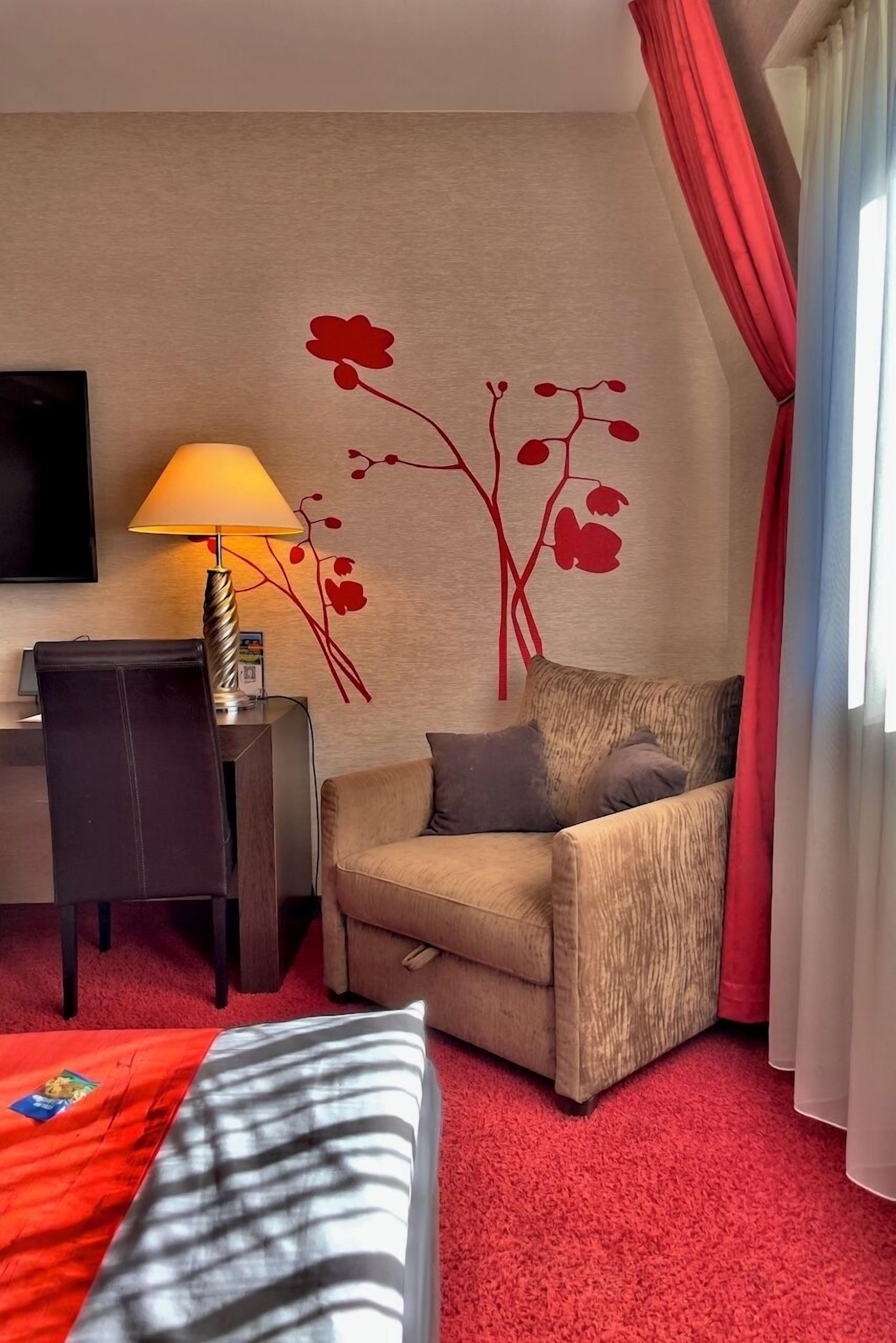 A hotel room with a sofa, a desk, a lamp, a TV, and a red carpet.