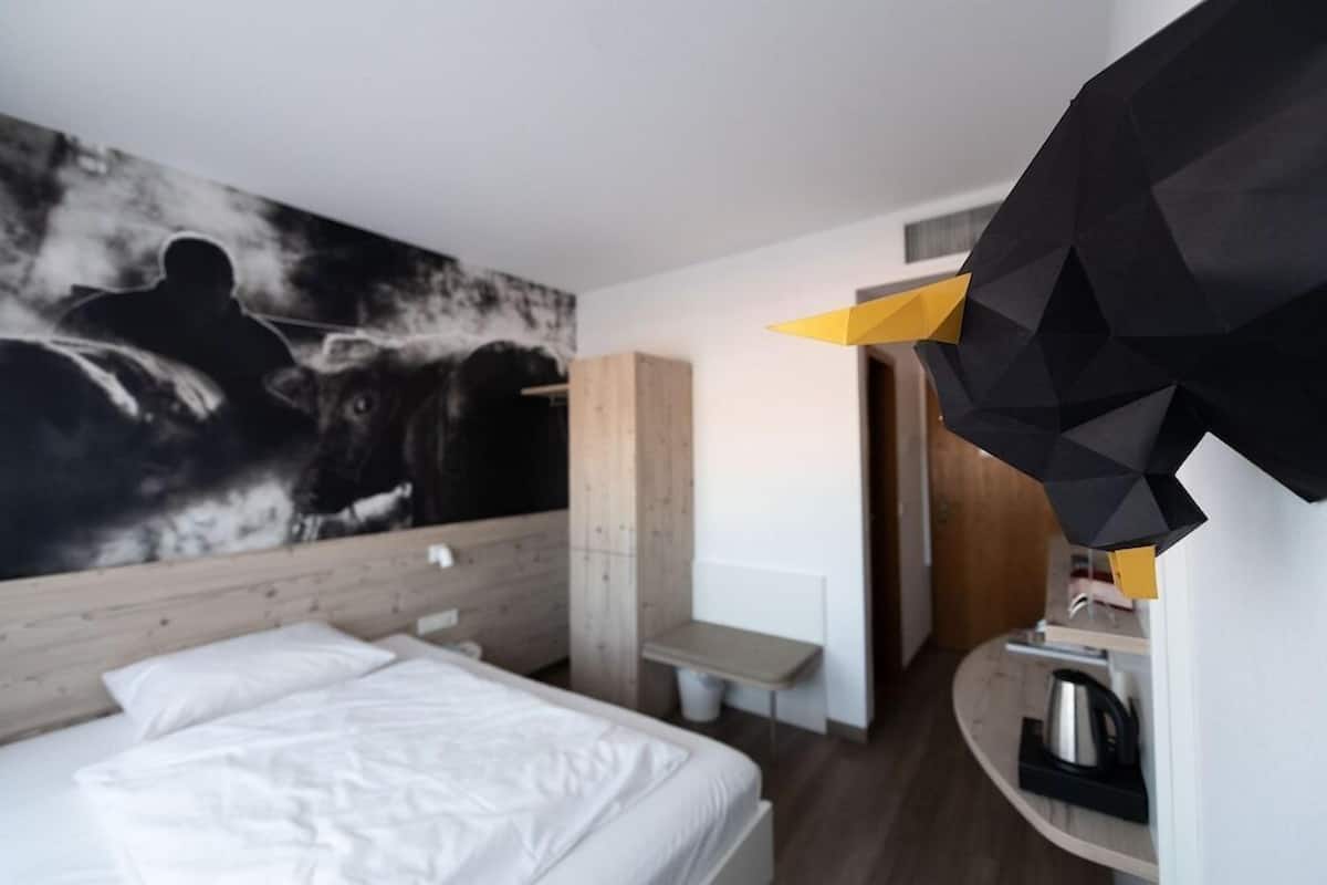 A modern hotel room with a large bed, a wooden headboard, a wall-mounted artwork, a small table, and a kitchen area with a kettle.