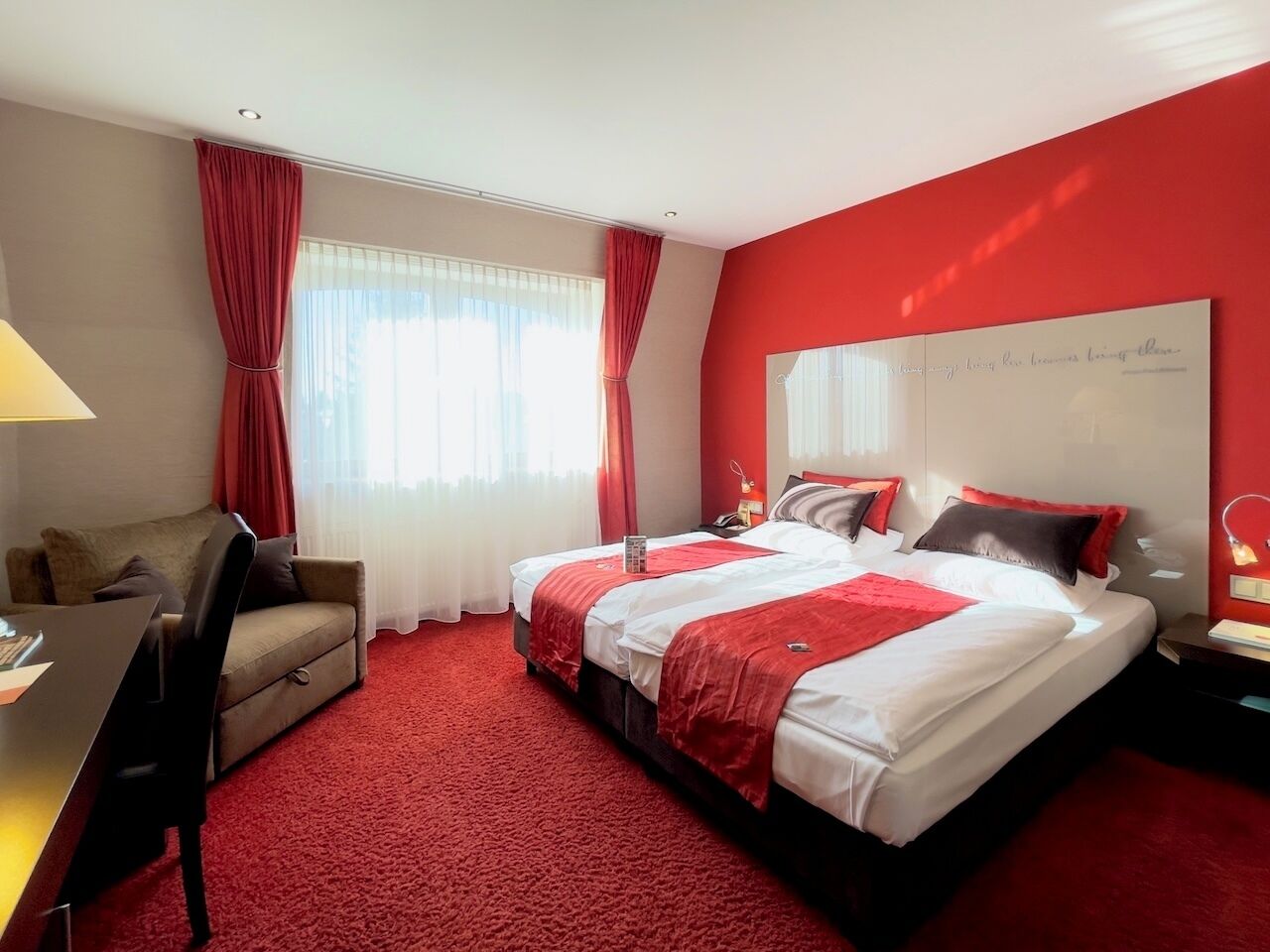 A hotel room with a red carpet, two beds, a desk, and a chair.