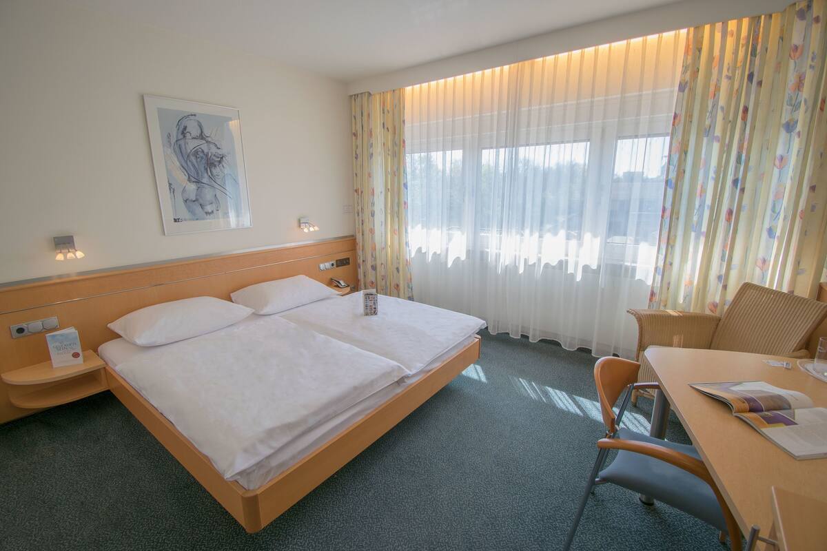 A hotel room with a double bed, a desk with a chair, a small table, a telephone, and a framed picture on the wall.