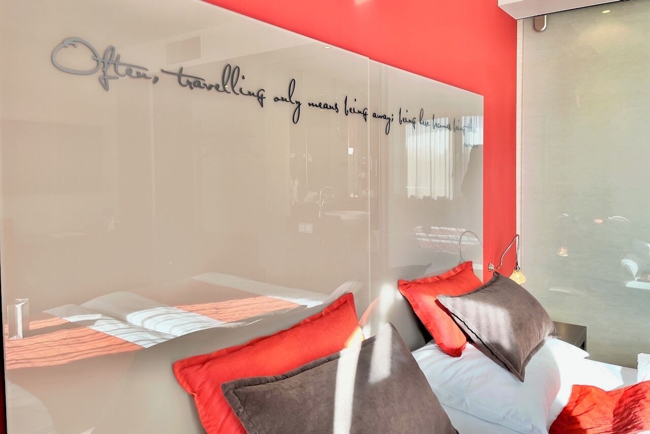 A modern hotel room with a large mirror, a bed with red and gray pillows, and a motivational quote on the mirror.