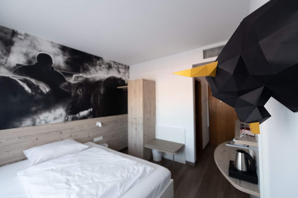 A modern hotel room with a large bed, a wooden headboard, a wall-mounted artwork, a desk, and a coffee machine.
