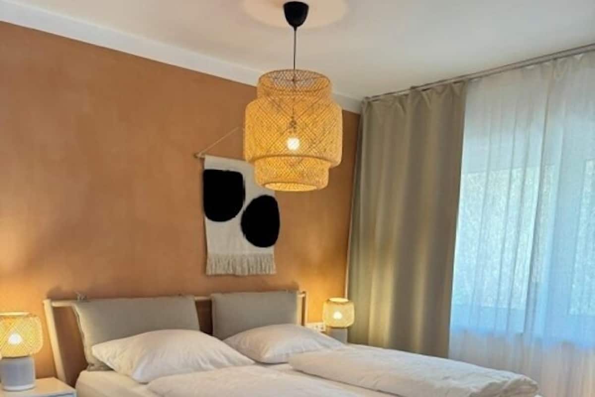 A bedroom with a bed, bedside tables, a hanging light fixture, and a window with curtains.