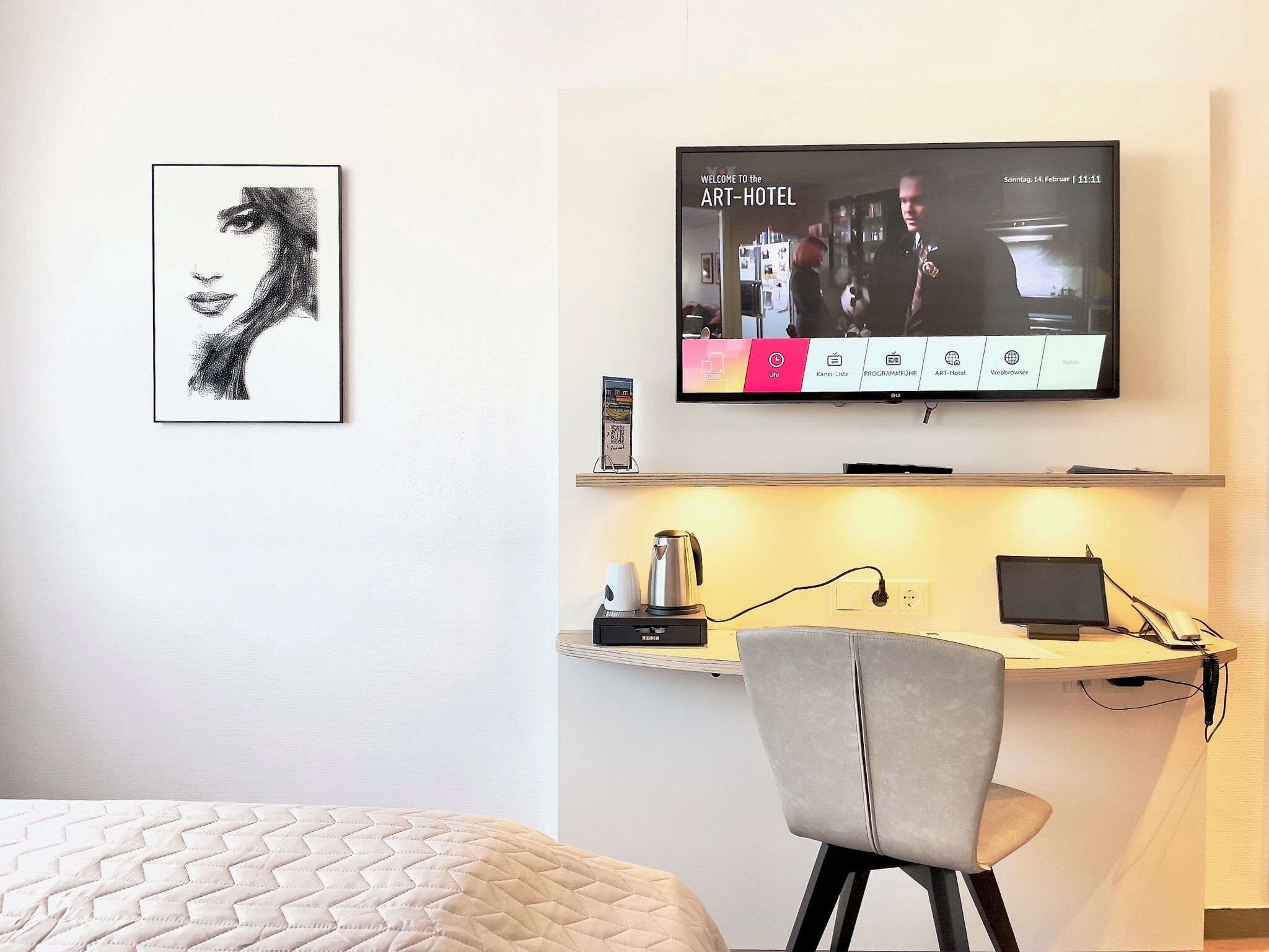 A hotel room with a bed, a desk with a laptop, a television displaying the Art-Hotel, and a coffee machine.