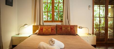 Una camera d'albergo con un letto grande, due cuscini, una finestra con tende, un comodino con una lampada e un segnale di uscita.