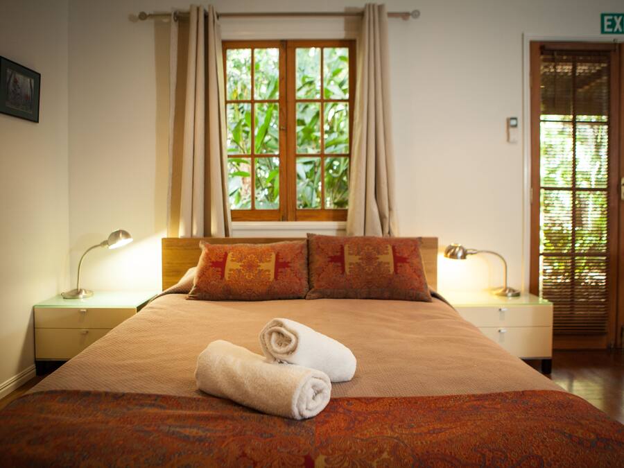 Una habitación de hotel con una cama grande, dos almohadas, una ventana con cortinas, una mesita de noche con lámpara y una señal de salida.