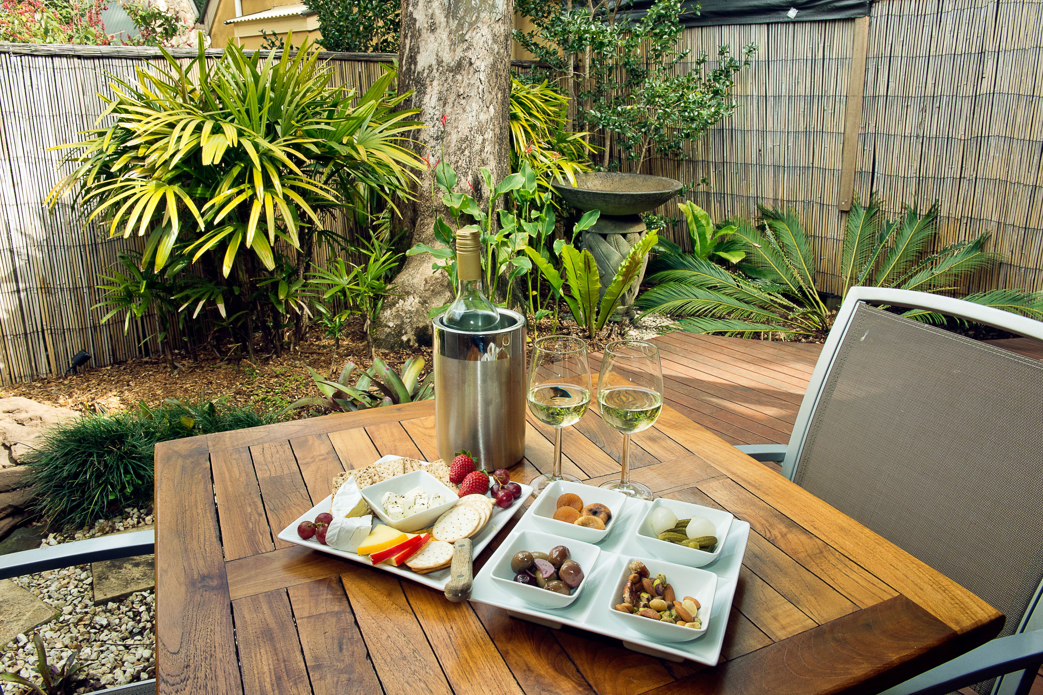 A wooden table set with a cheese board, fruits, nuts, and two glasses of white wine.