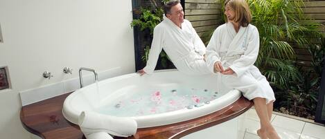 A couple in white bathrobes sitting in a freestanding bathtub with a wooden edge, surrounded by greenery.