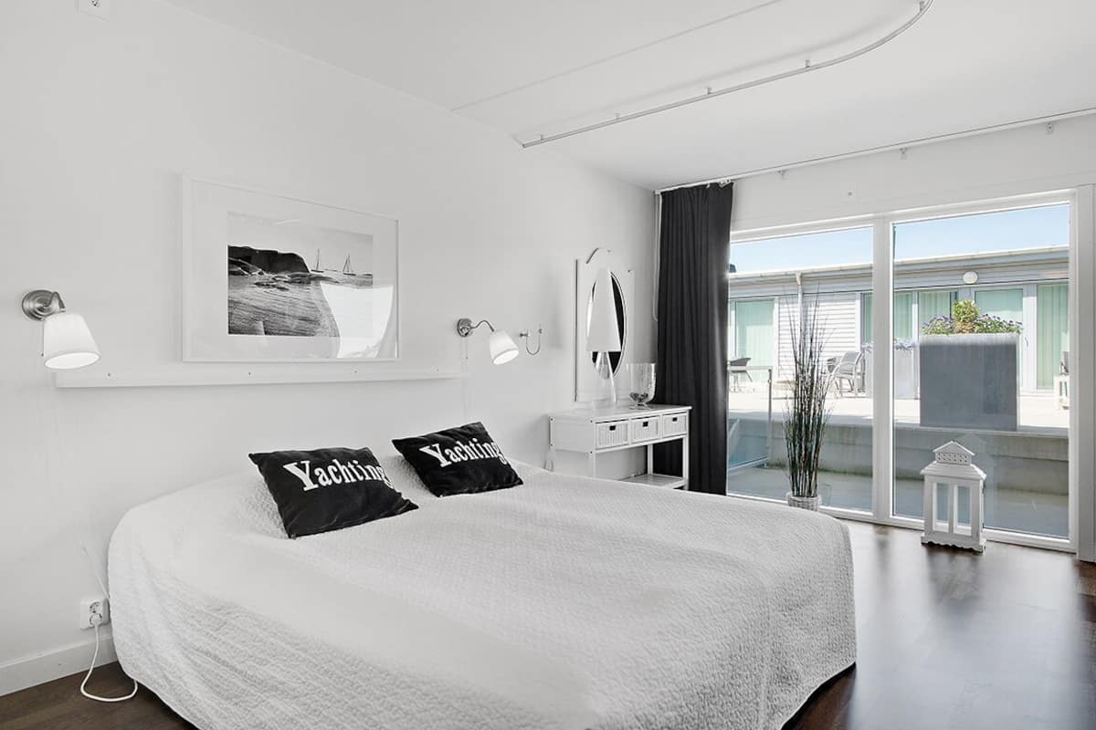A modern bedroom with a large bed, two black pillows with text, a bedside table, a mirror, and a view of the outside through a sliding glass door.