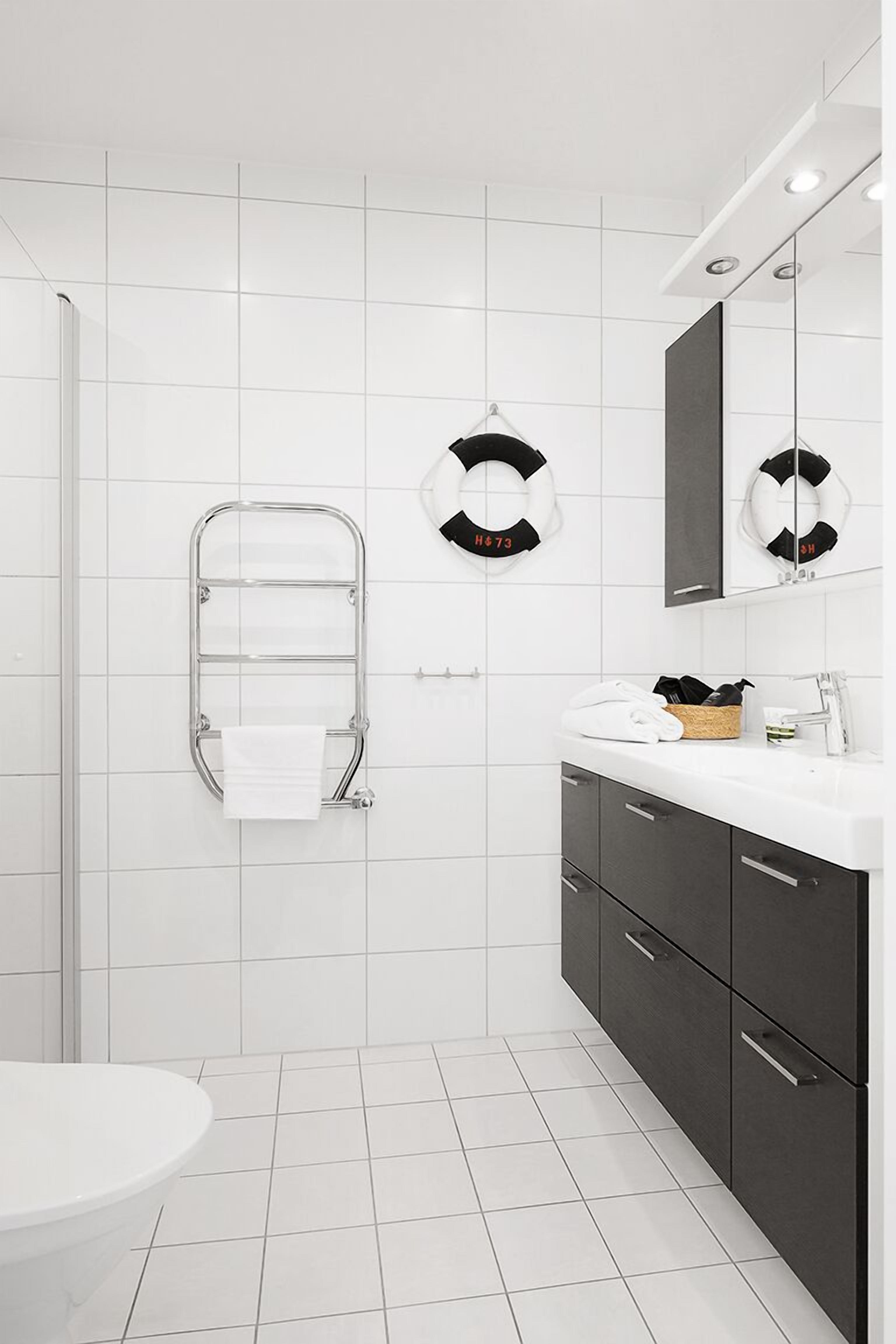 A modern bathroom with white tiles, a glass shower enclosure, a towel rack, a sink with a mirror, and dark cabinets.
