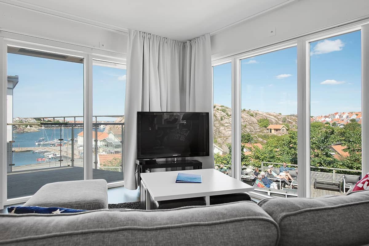 A modern living room with a large TV, a white coffee table, and a sofa. It features large windows with a view of a marina and buildings.