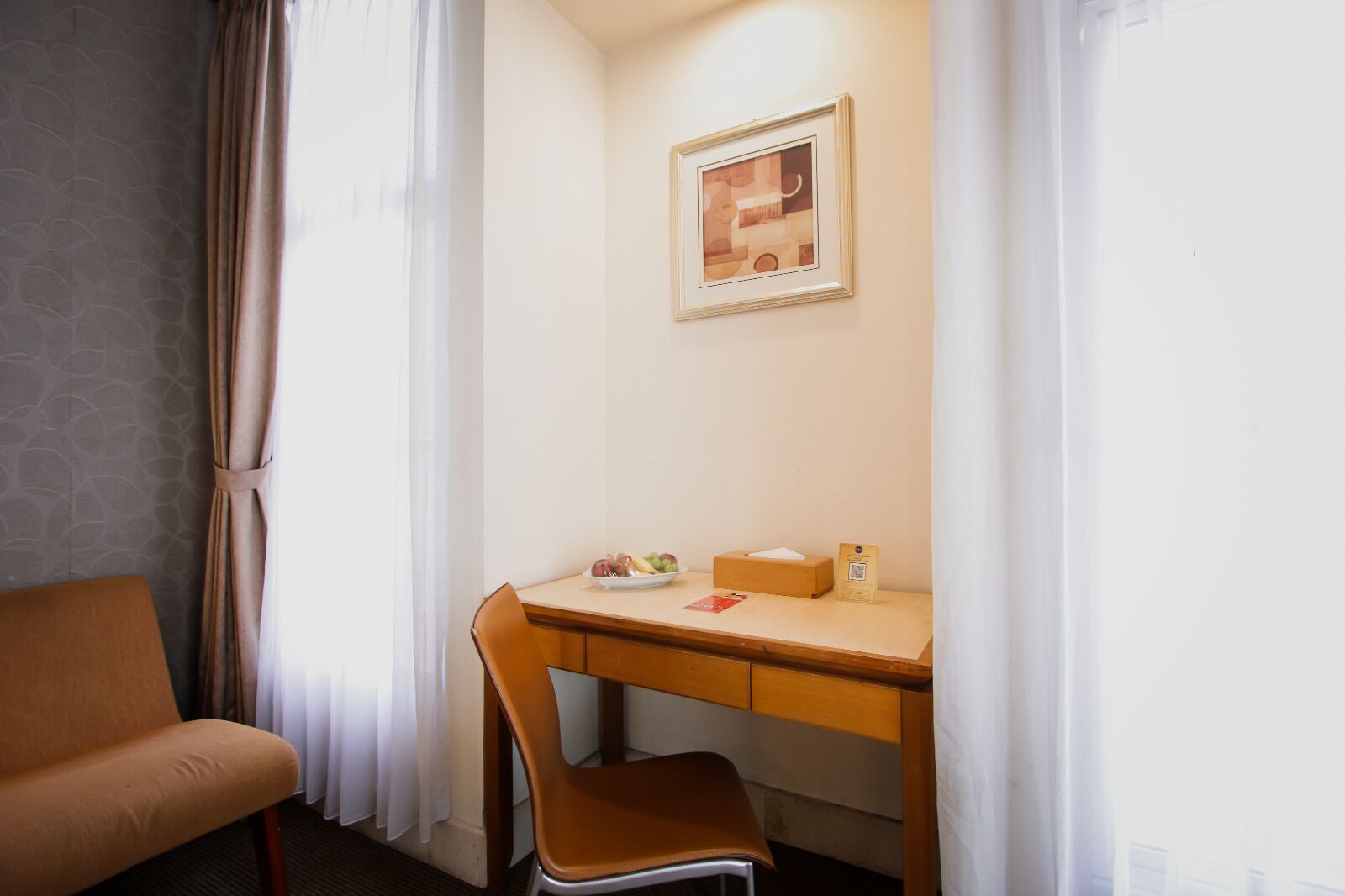 A corner room with a wooden desk, a chair, a framed picture on the wall, and a window with curtains.