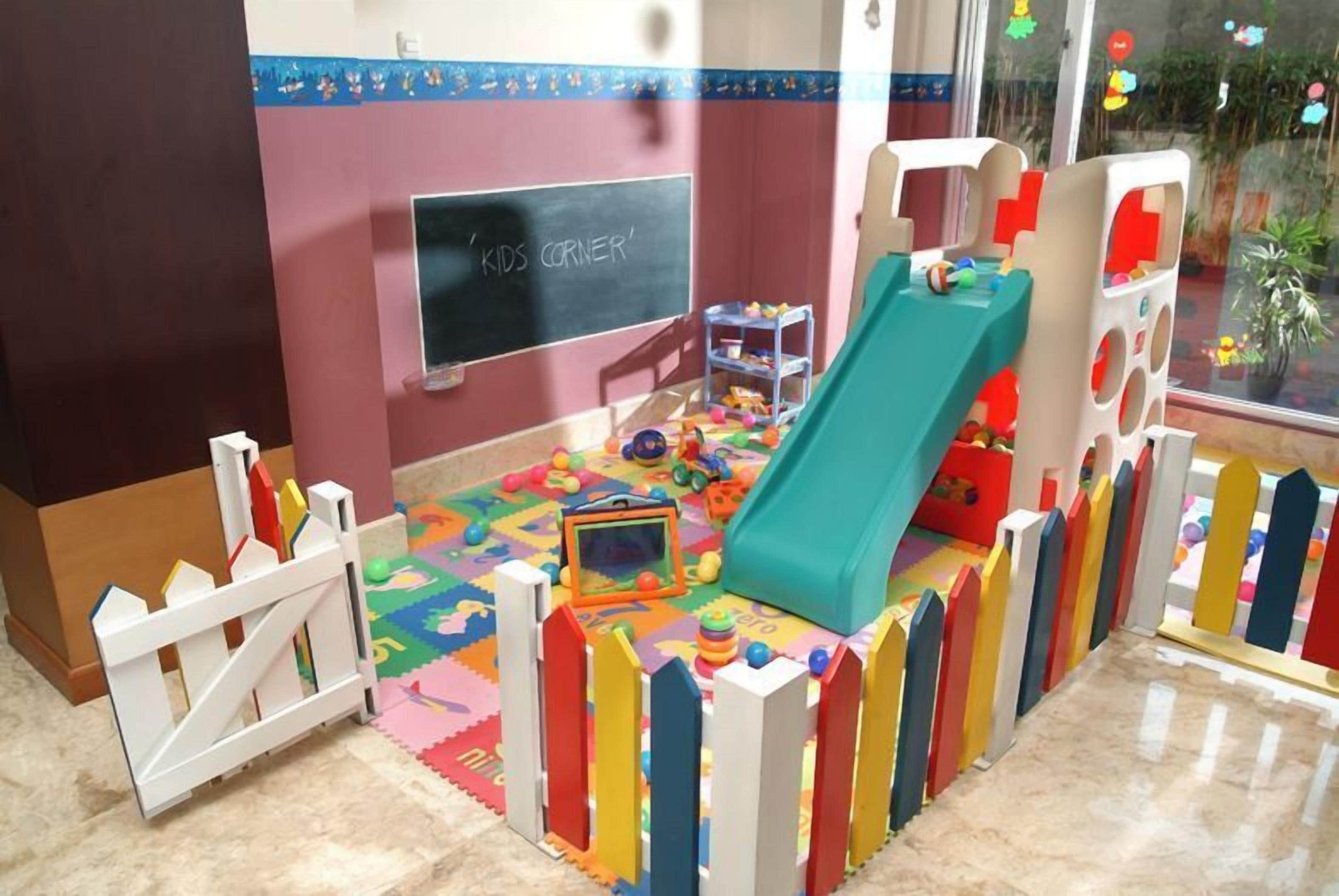 Espaço infantil com escorregador, tapetes de brincadeira coloridos e um quadro negro com a inscrição 'KIDS CORNER'.
