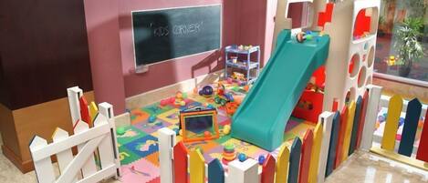 Espaço infantil com escorregador, tapetes de brincadeira coloridos e um quadro negro com a inscrição 'KIDS CORNER'.