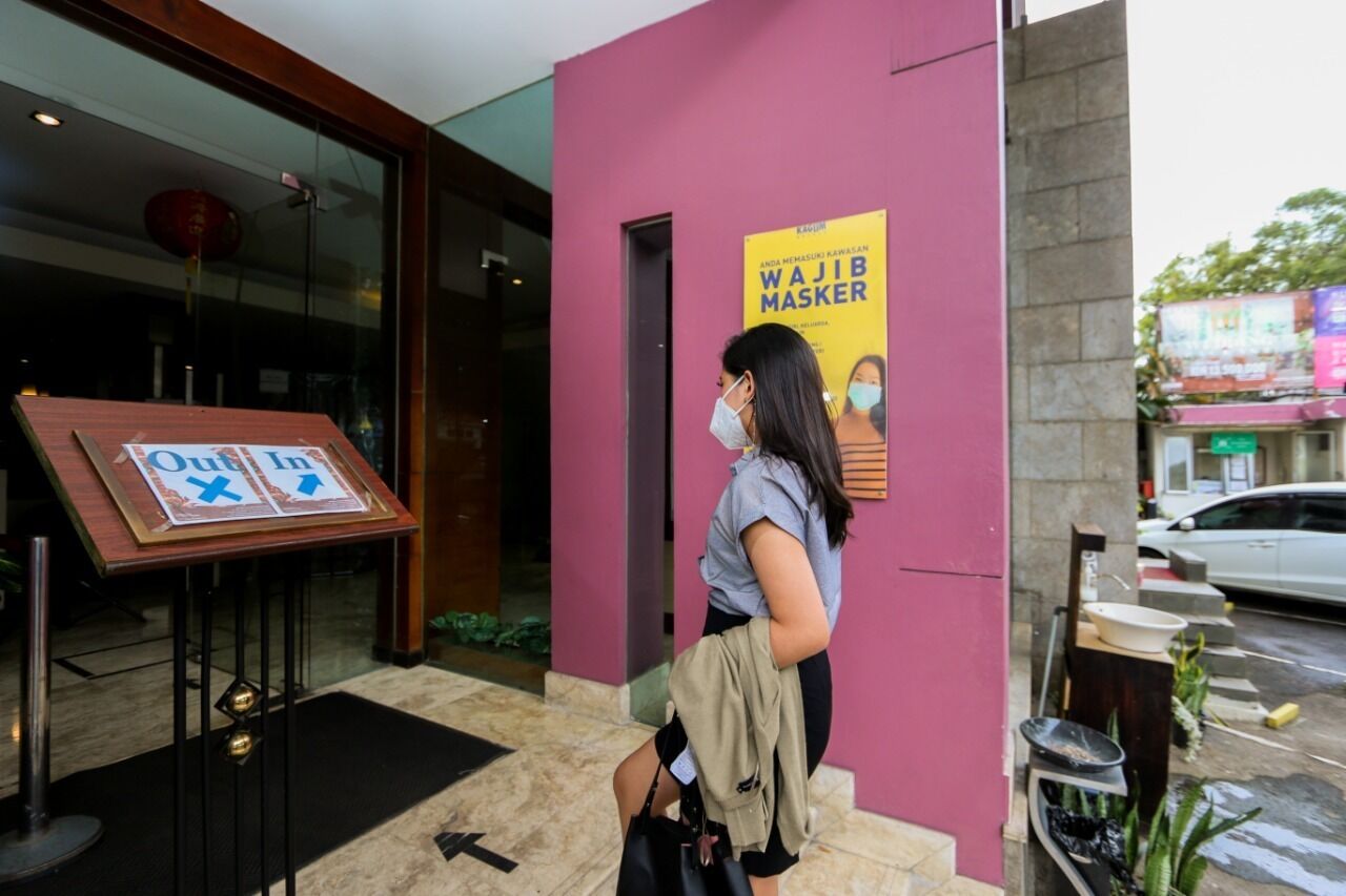 Une femme se tient à l’entrée d’un bâtiment, devant une enseigne indiquant « Entrée/Sortie » et une autre faisant la publicité d’un « Wajib Masker ».
