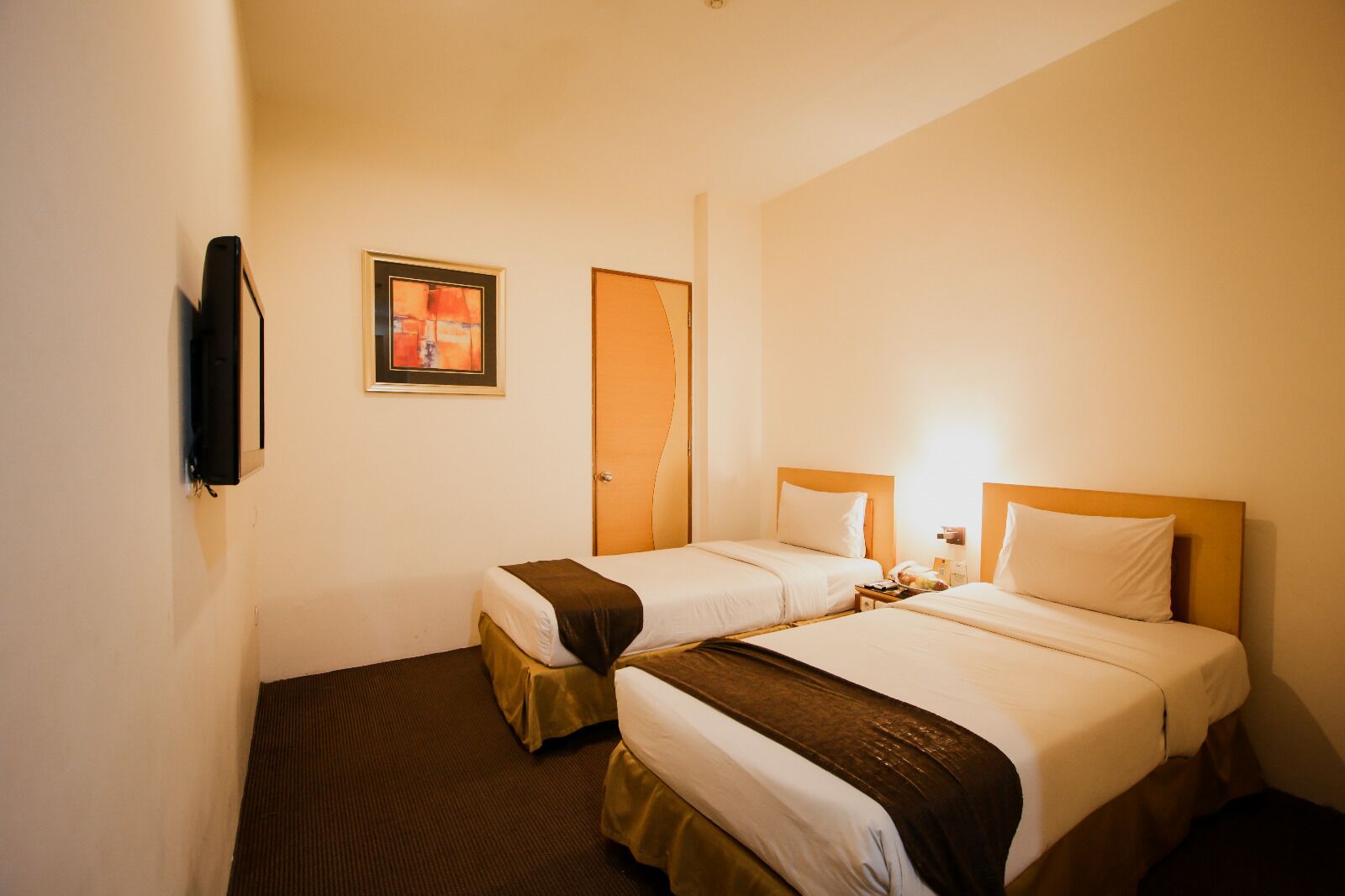 A hotel room with two beds, a door, a framed picture, and a flat-screen TV mounted on the wall.