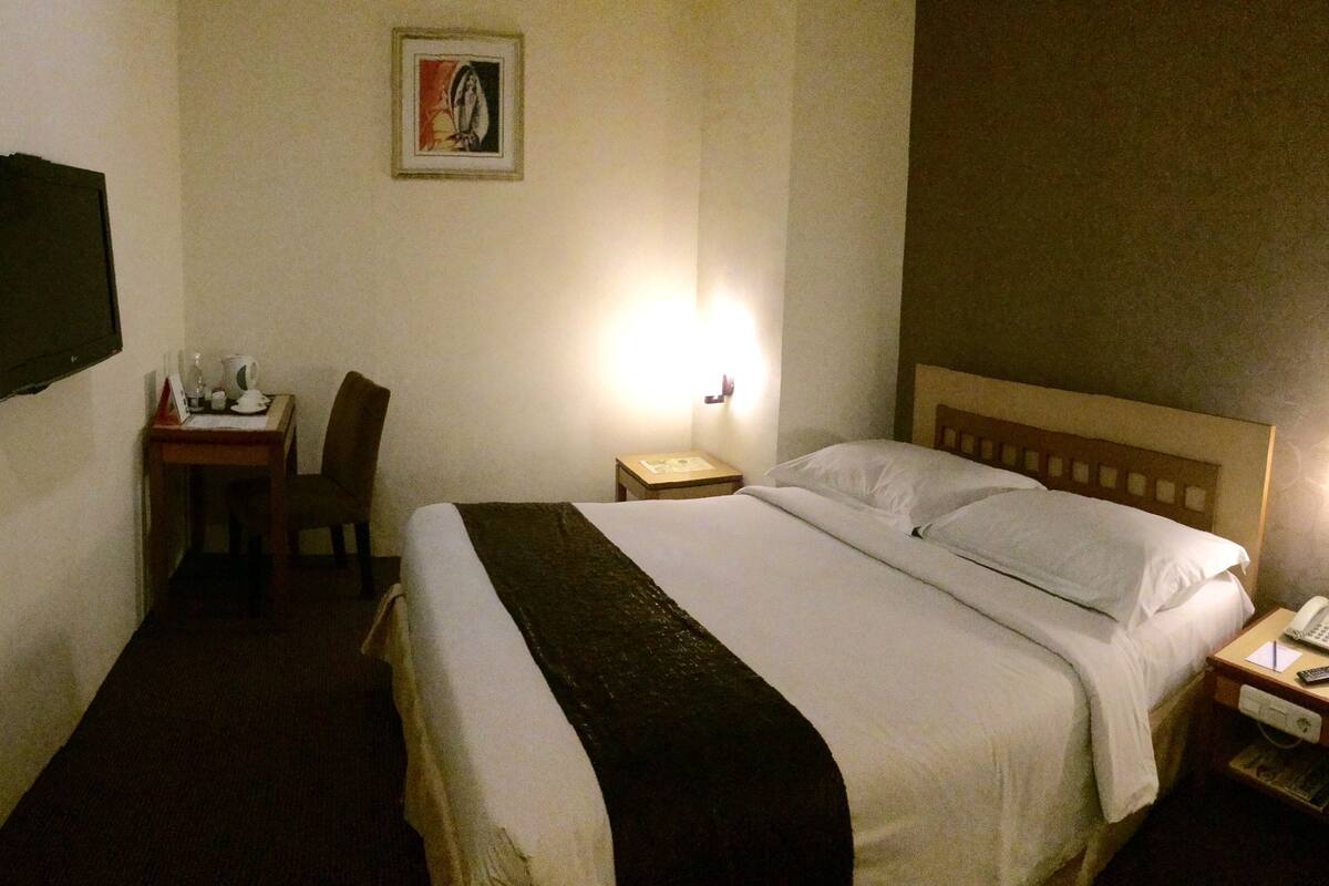 A hotel room with a bed, bedside table with a phone and remote, a desk with a chair, a wall-mounted TV, and a painting on the wall.