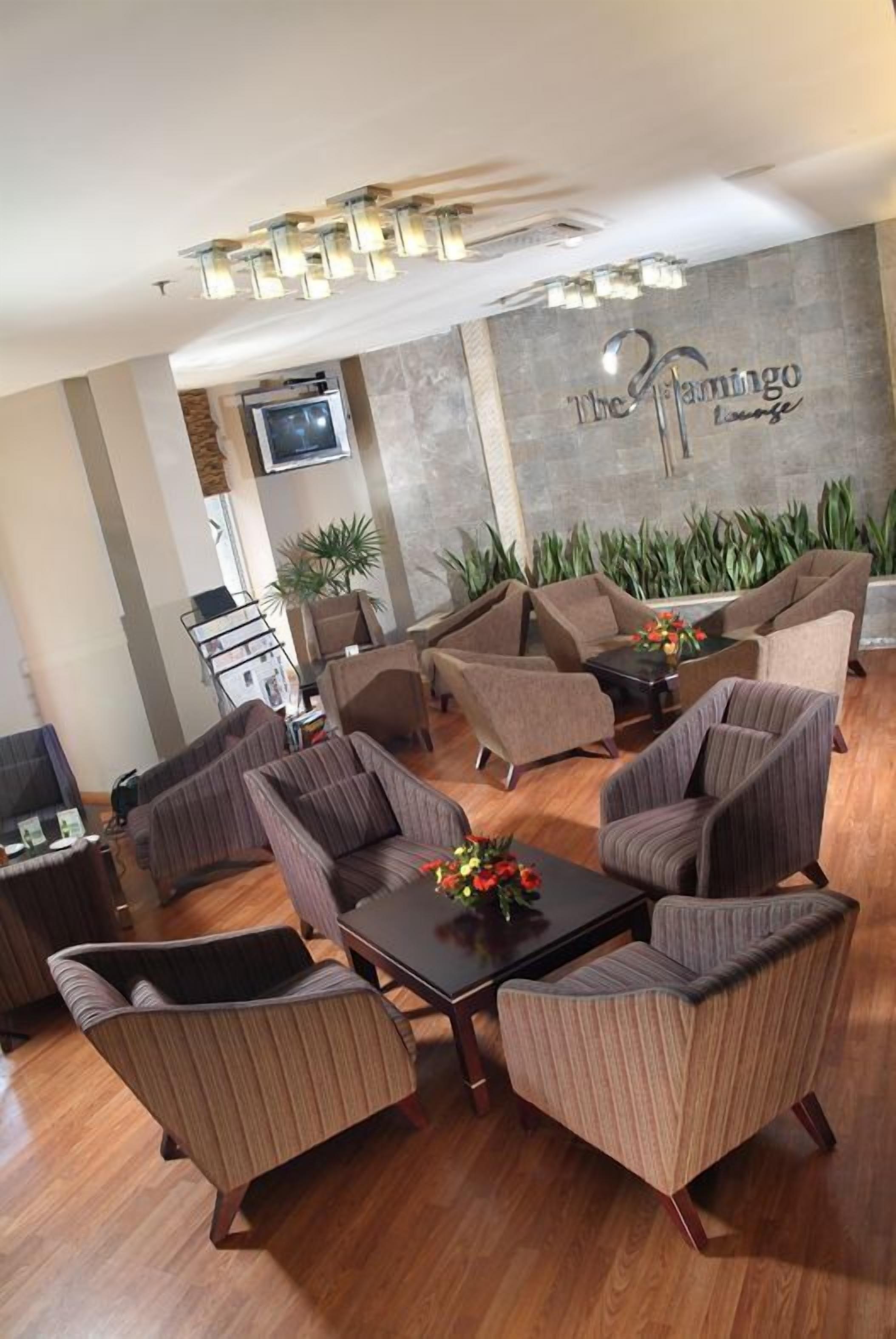 Un espace détente avec plusieurs canapés et fauteuils, une table basse agrémentée de fleurs, un téléviseur à écran plat fixé au mur, et un mur décoratif portant l’inscription « The Flamingo Lounge ».