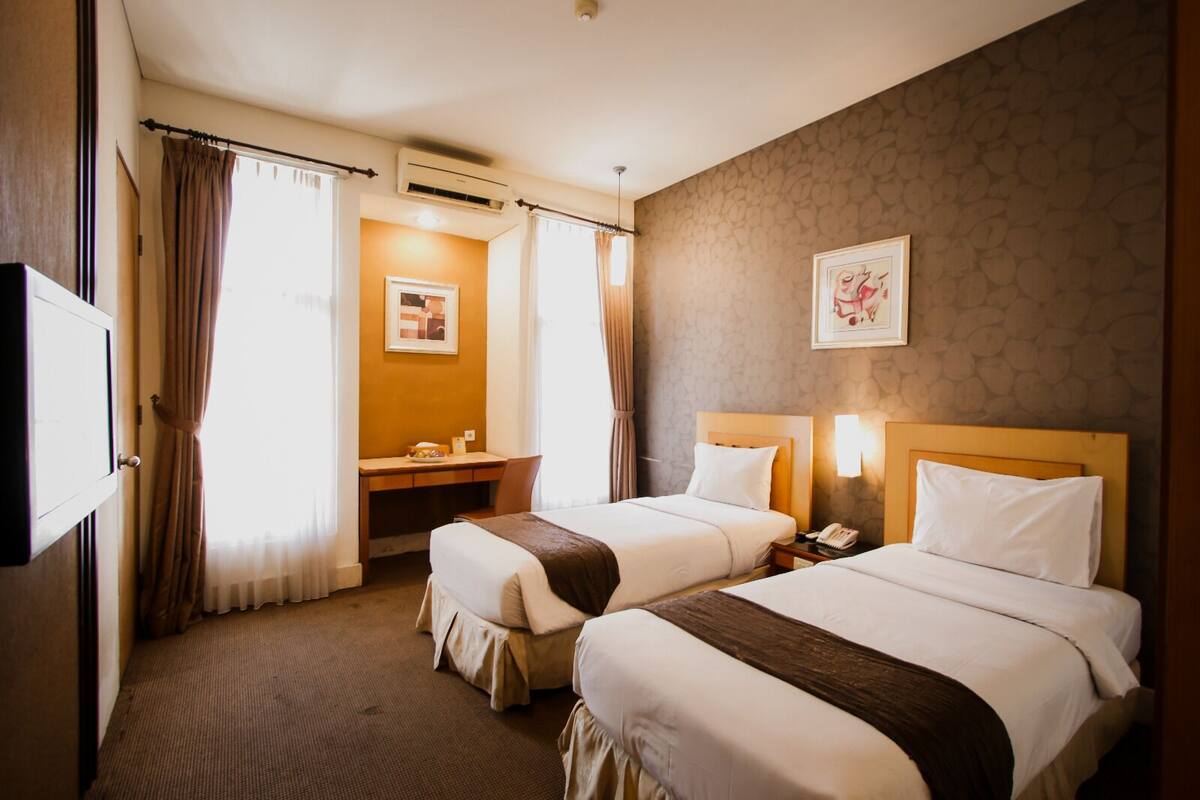 A hotel room with two beds, a desk, a chair, an air conditioning unit, a telephone, and a framed picture on the wall.
