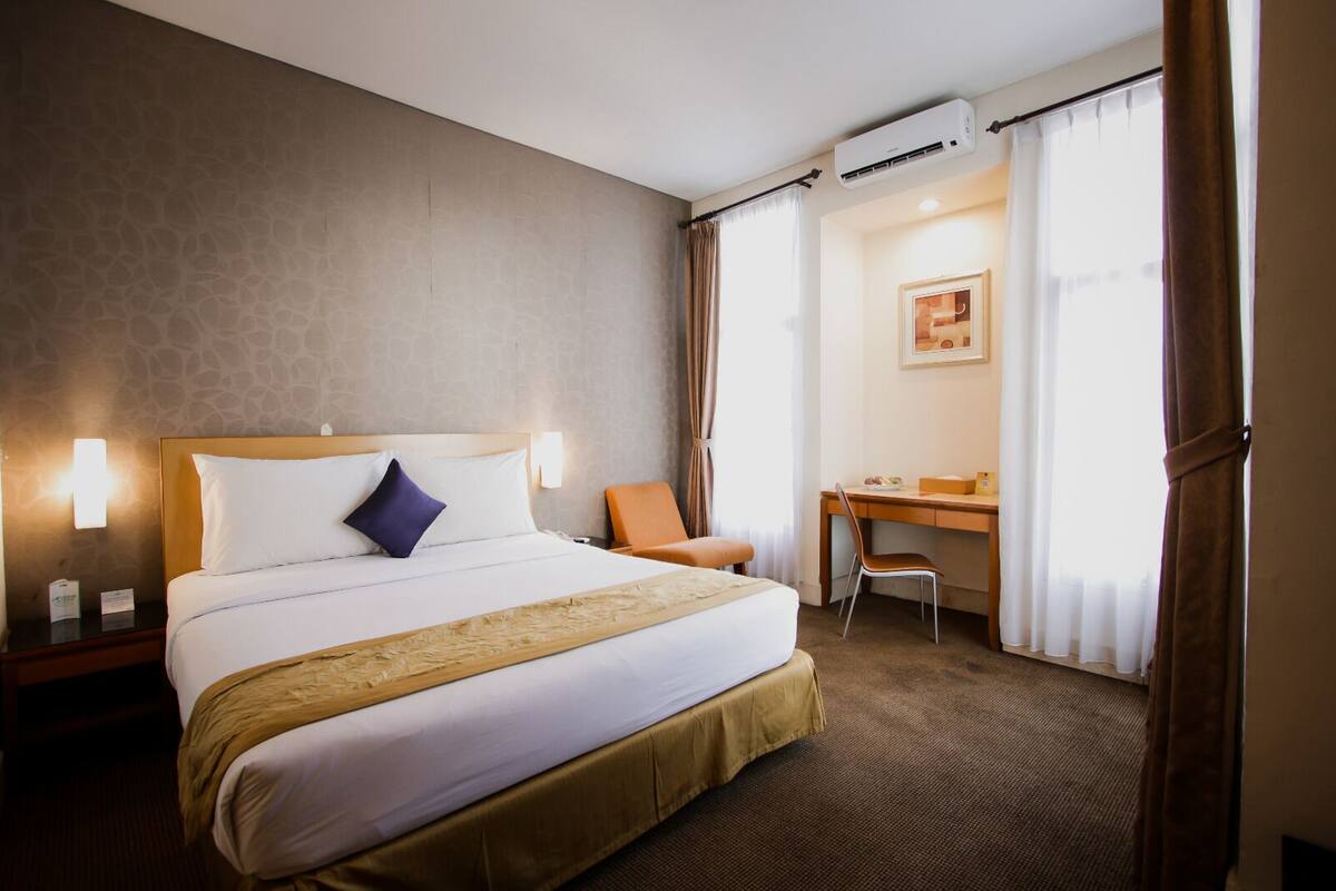 A hotel room with a bed, a desk, a chair, an air conditioning unit, and a window with curtains.
