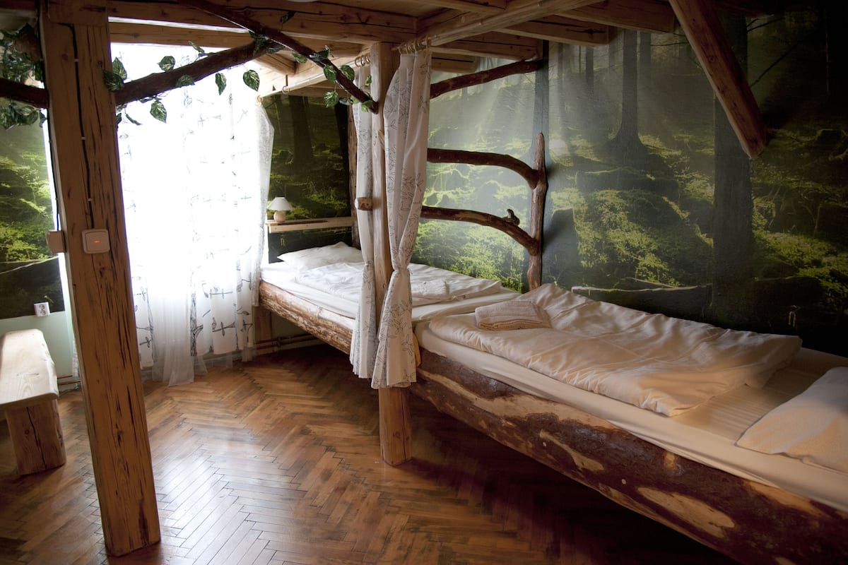 Una habitación rústica de madera con una cama, un banco y vistas a un bosque a través de una ventana.