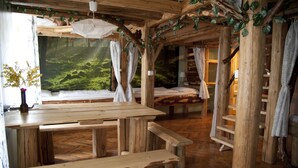 A rustic wooden cabin interior with a large bed, a wooden table, benches, and a view of a forest through a large window. - Artharmony Pension & Hostel (Prague)