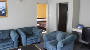 Ein Hotelzimmer mit zwei blauen Sesseln, einem schwarzen Couchtisch, einem Radio auf einem Regal und einem Bett mit brauner Kopfteil im Hintergrund.
