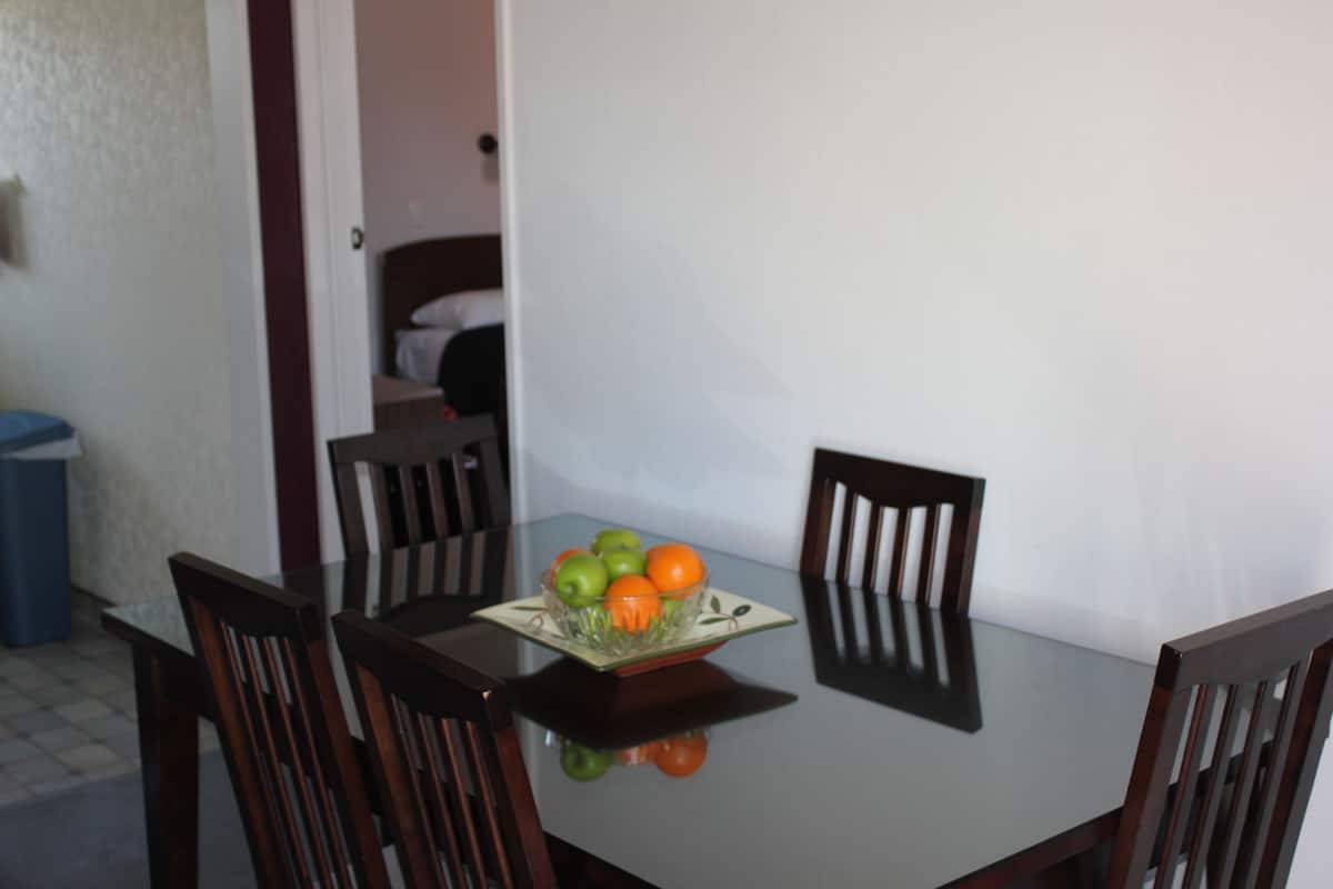 Une table à manger avec des chaises, un saladier en verre rempli de fruits et un reflet sur la surface de la table.
