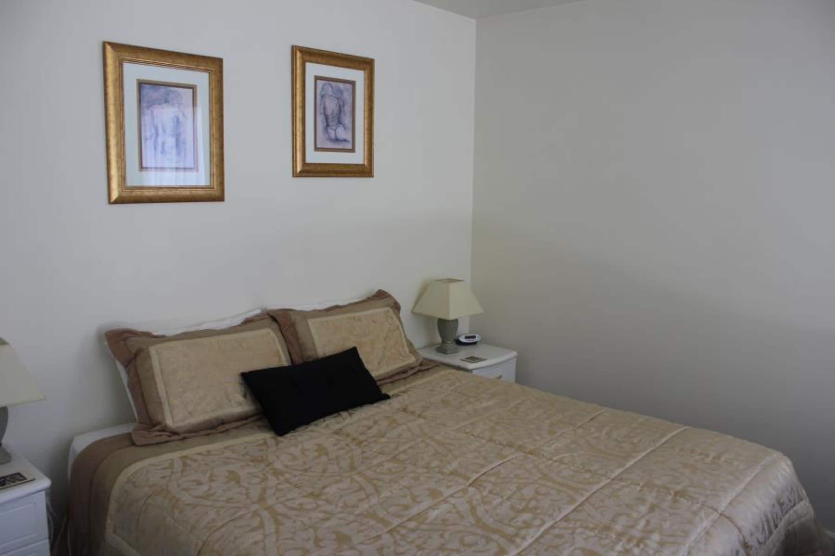 Una cama bien hecha con un edredón estampado, dos cuadros en la pared y una lámpara de noche sobre una mesita blanca.