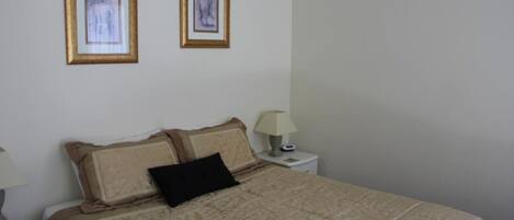 Una cama bien hecha con un edredón estampado, dos cuadros en la pared y una lámpara de noche sobre una mesita blanca.