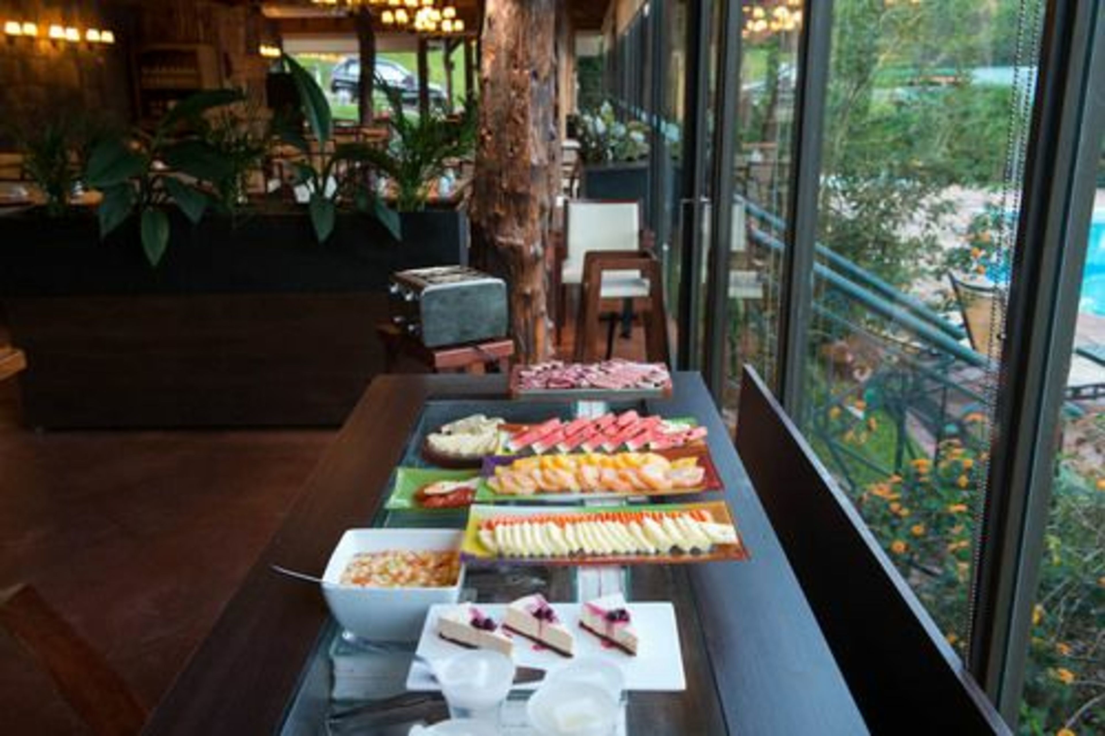 A buffet table with various food items and a view of a garden through large windows.