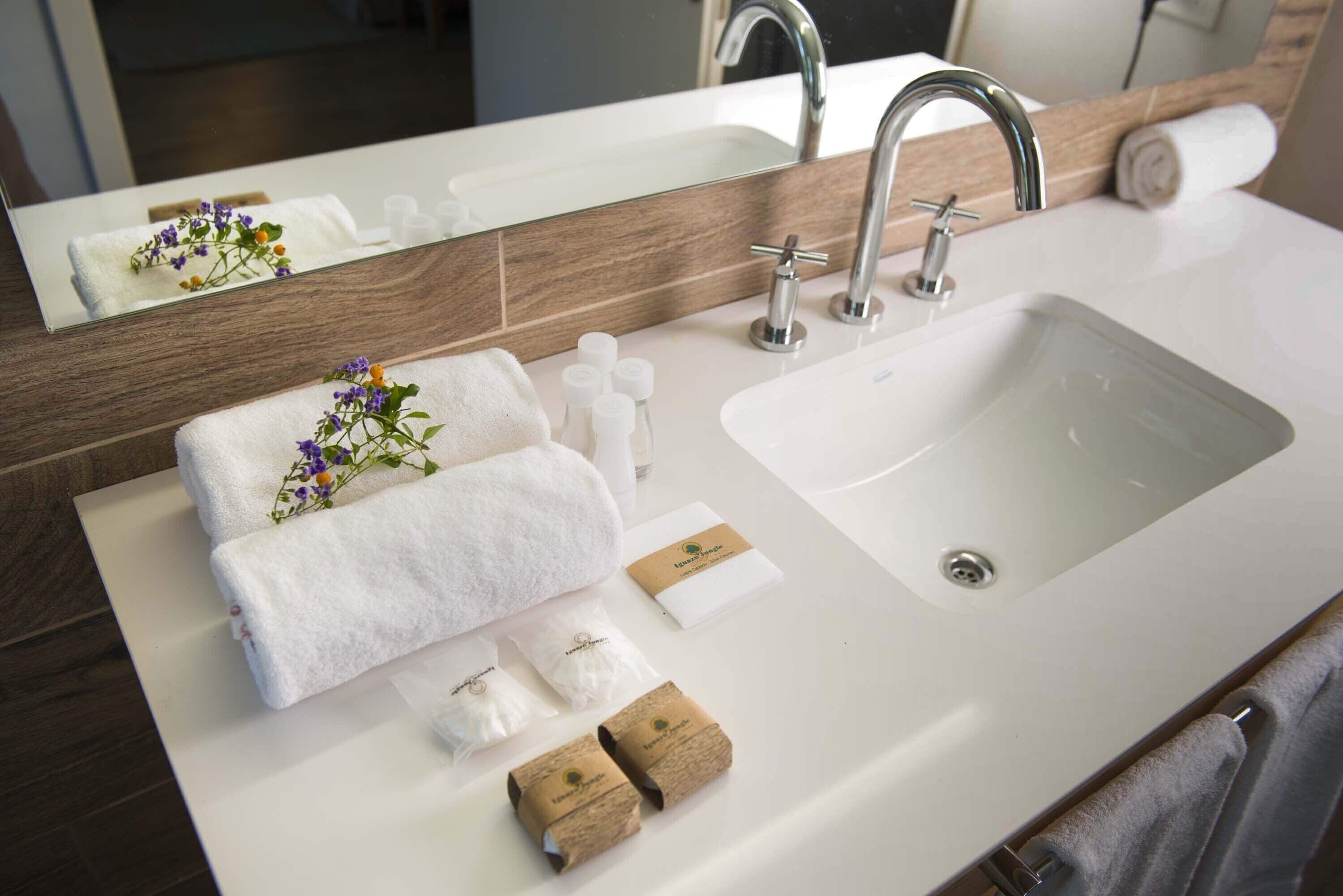 A bathroom with a white sink, two faucets, a mirror, and toiletries including a towel, soap, and a small bottle.