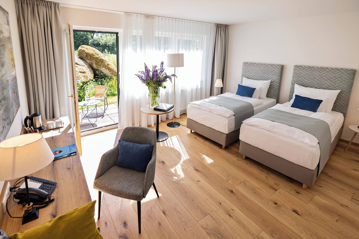A hotel room with two beds, a chair, a desk with a lamp, and a view of the outdoors.