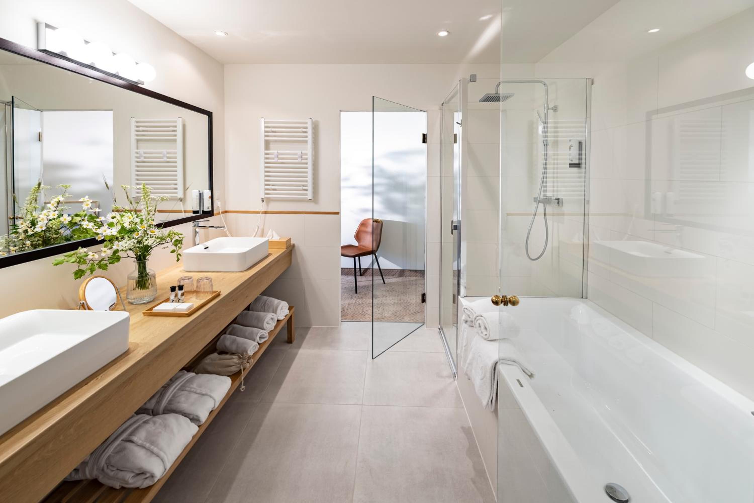 A modern bathroom with a large mirror, two sinks, a shower, and a bench with towels.